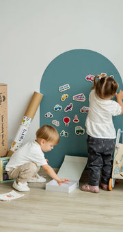 Two toddlers playing with Tix&Mix wooden magnetic vehicles playset on a cyan wave magnetic wall decal.