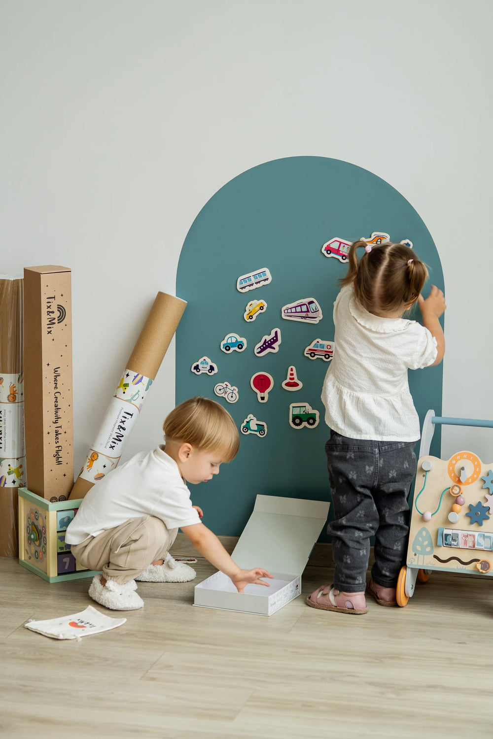 Two toddlers playing with Tix&Mix wooden magnetic vehicles playset on a cyan wave magnetic wall decal.