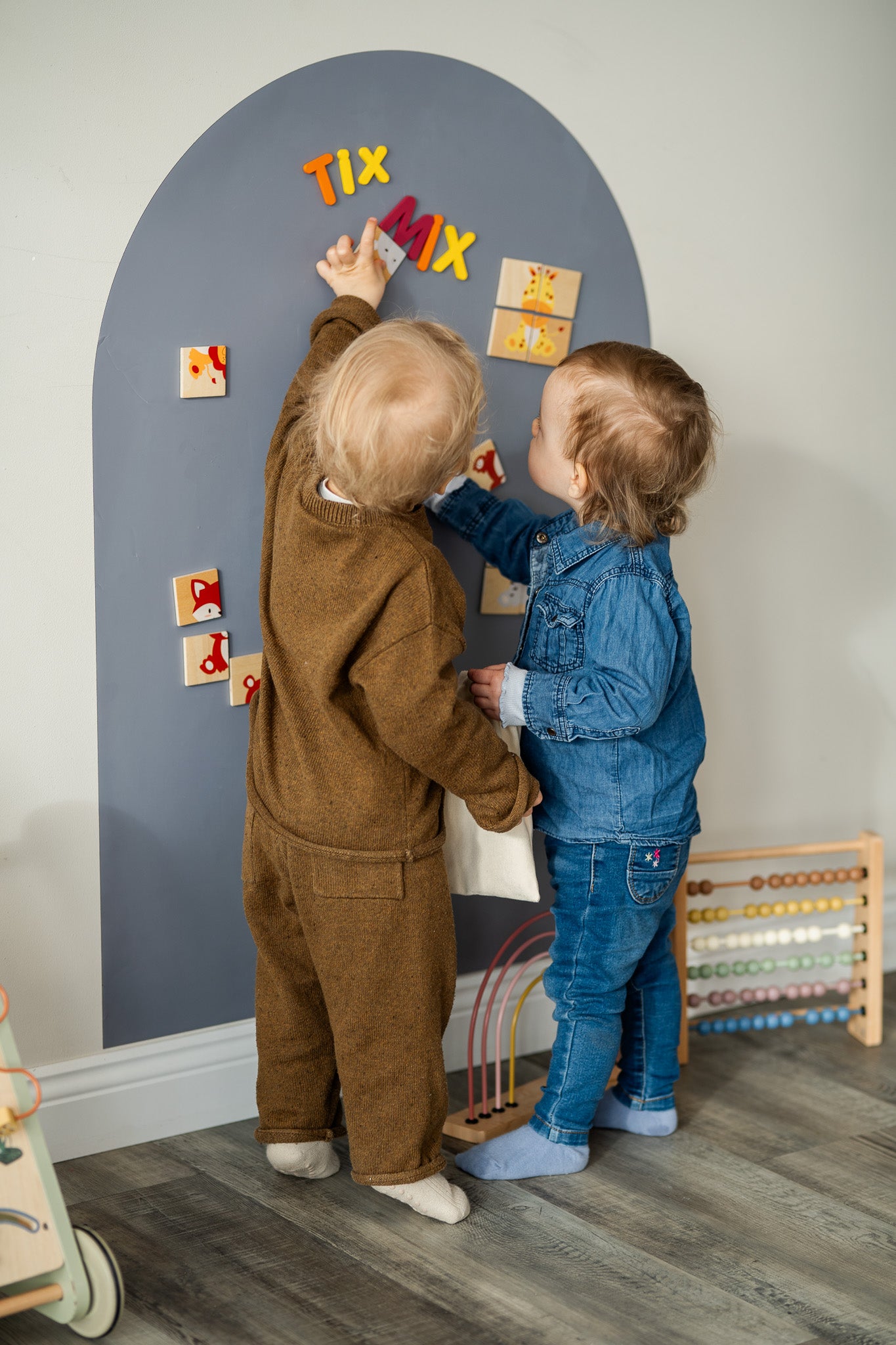 Two toddlers playing with Tix&Mix animals puzzle play set on a grey magnetic wall decal.