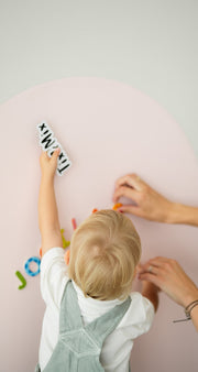 A toddler placing TixMix  letter pieces on a pink magnetic wall board while being assisted by a parent.