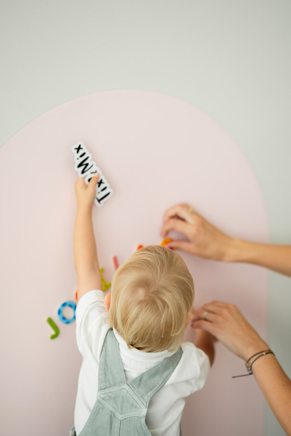 A toddler placing TixMix  letter pieces on a pink magnetic wall board while being assisted by a parent.