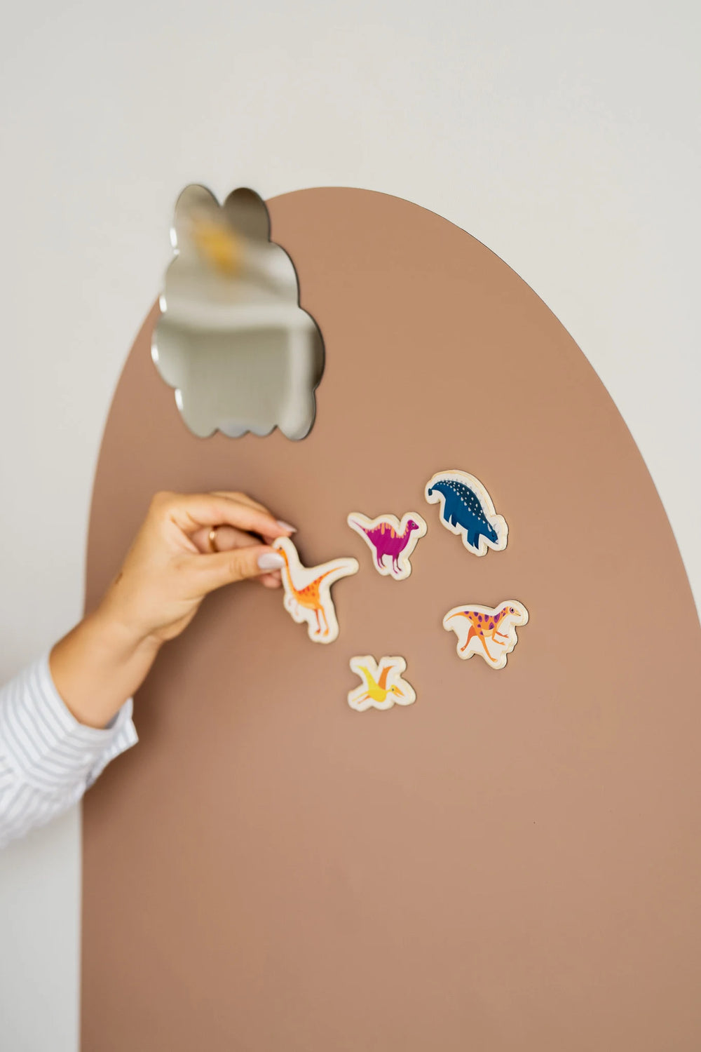 A hand placing colorful dinosaur toy on a brown arch-shaped magnetic wall decal featuring other dinosaur toys and a magnetic mirror.
