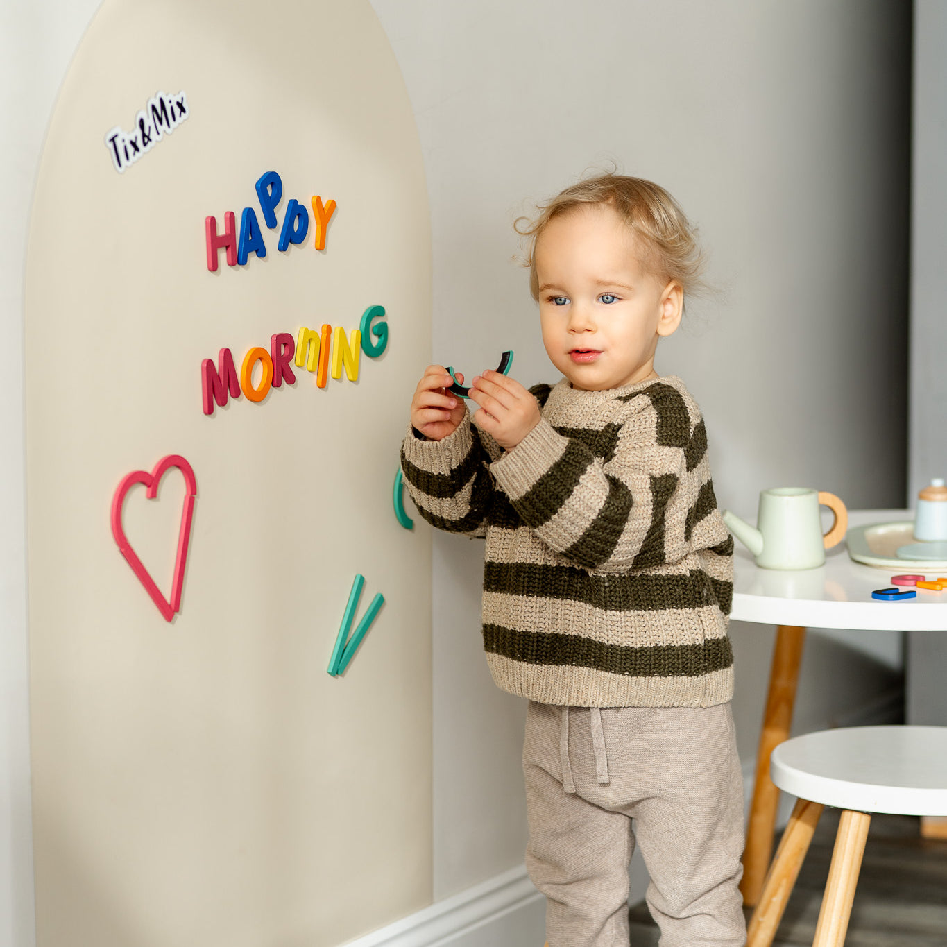 A toddler holding Tix&Mix rings and blocks play set with white magnetic wall decal on the wall.