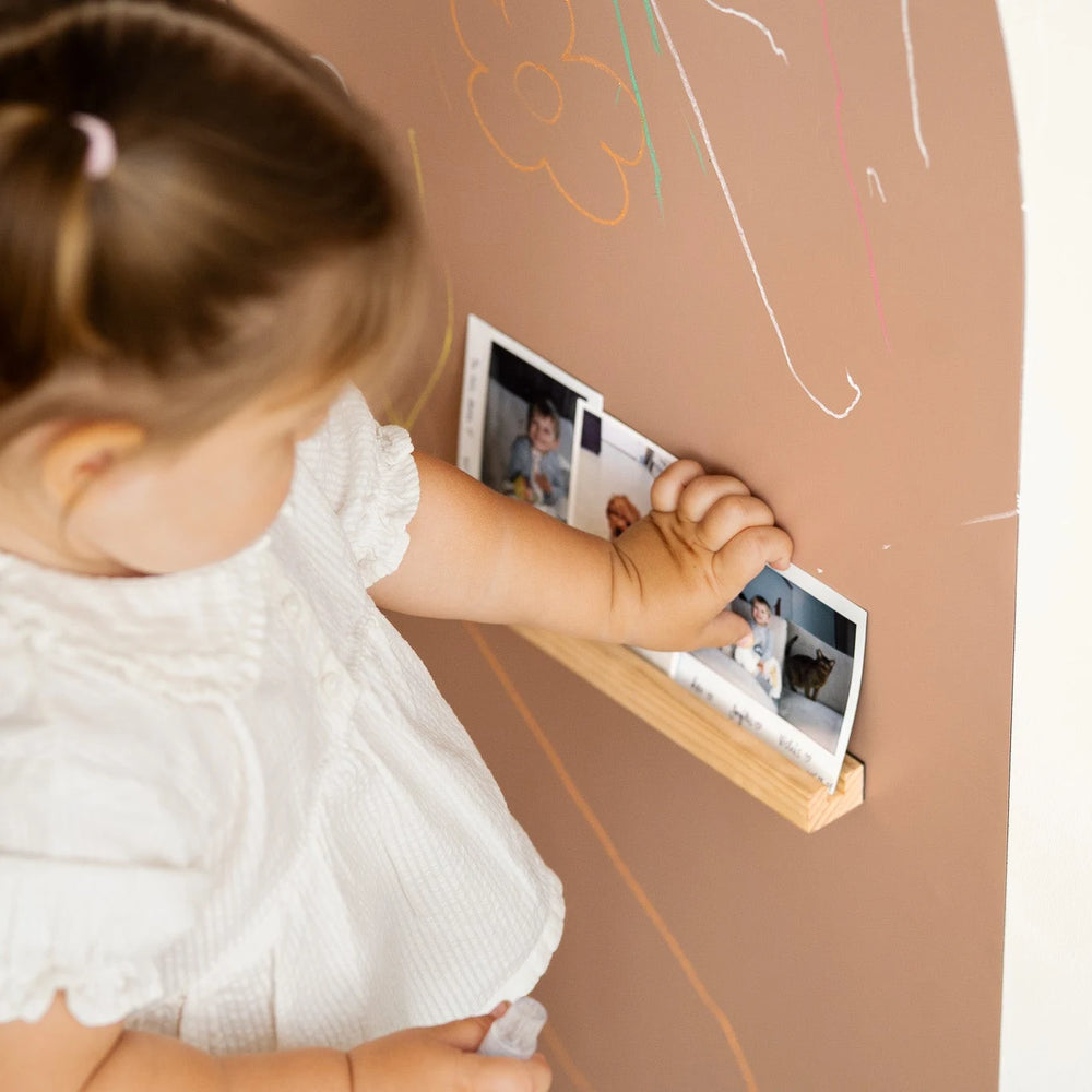 A female toddler placing printed pictures in a Tix&Mix magnetic wooden treasure shelf on a brown wall decal.