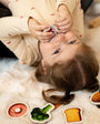A child laying on a clean floor with a magnetic toy on her mouth, and magnetic vegetable toys on the side.