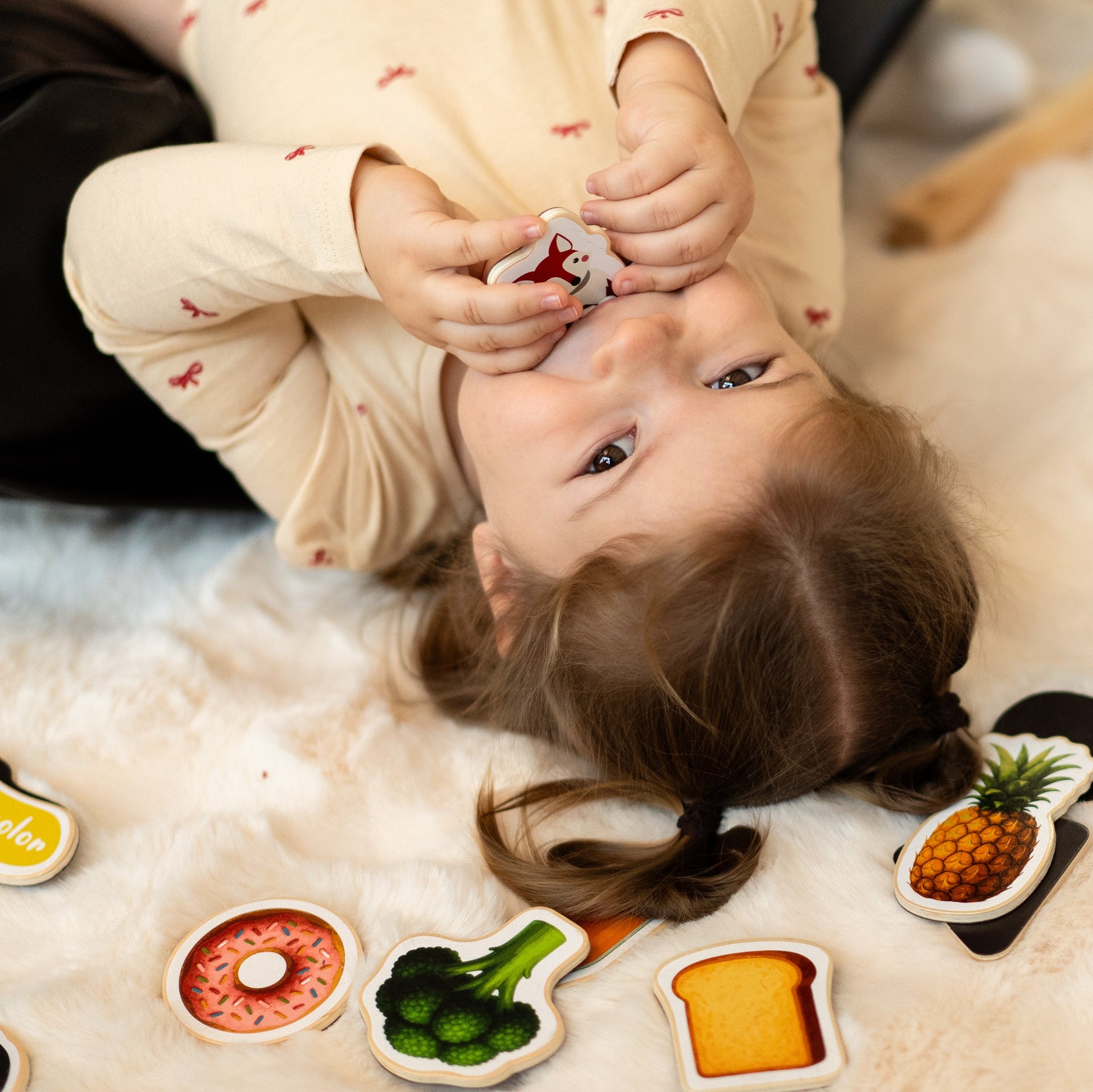 A child laying on a clean floor with a magnetic toy on her mouth, and magnetic vegetable toys on the side.