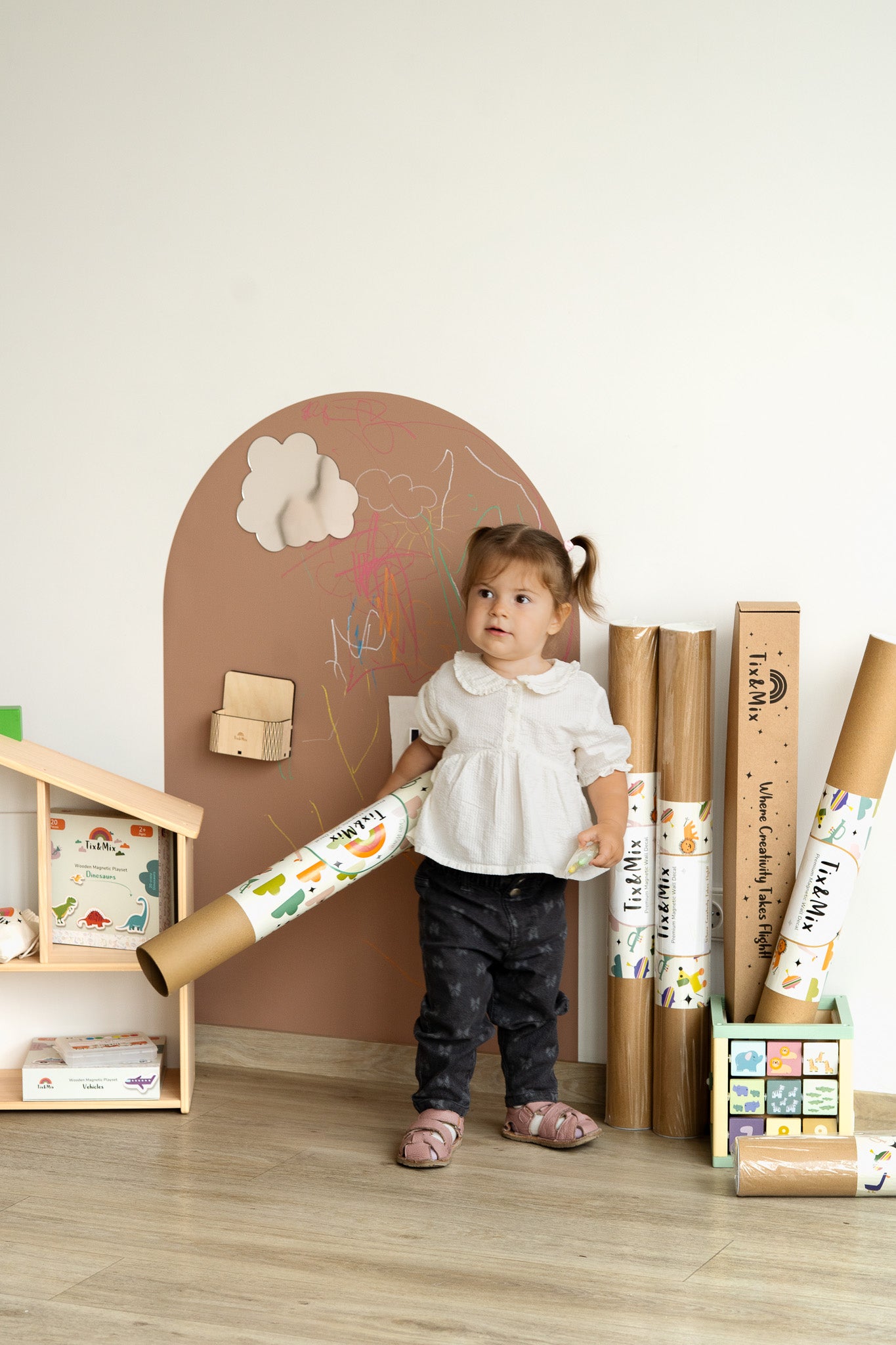 A toddler holding a Tix&Mix magnetic wall decal roll in a playroom, standing beside a brown wall board.