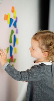 A girl toddler arranging colorful magnetic shapes vertically on a Tix&Mix wall decal.
