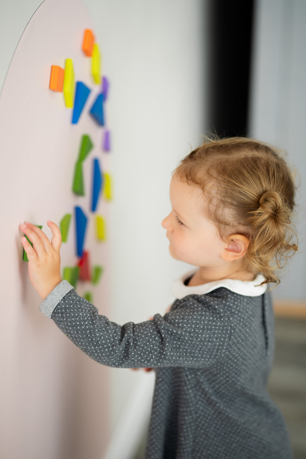 A girl toddler arranging colorful magnetic shapes vertically on a Tix&Mix wall decal.