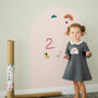 A toddler standing beside a Tix&Mix pink magnetic wall decal in a playroom.
