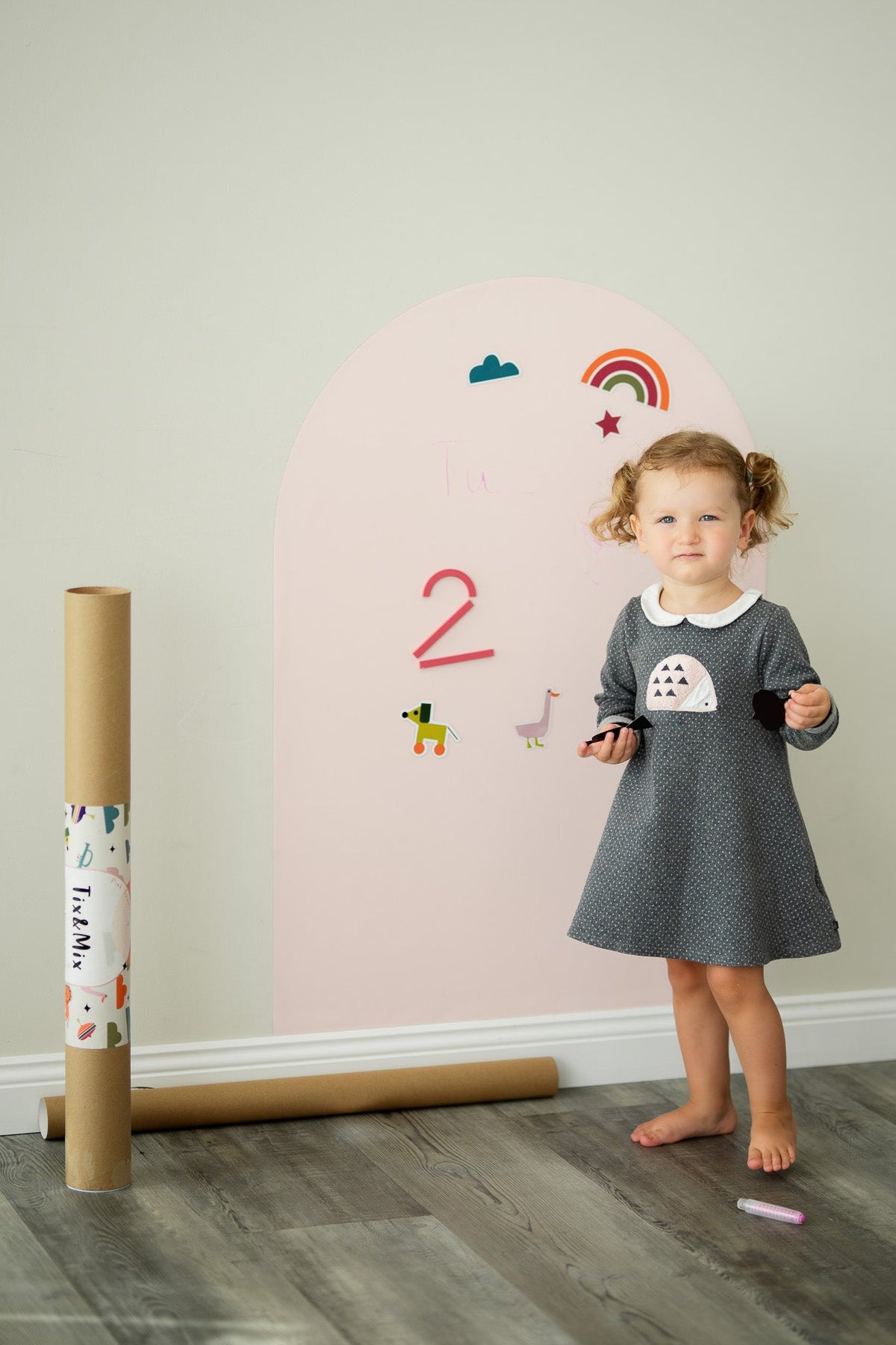 A toddler standing beside a Tix&Mix pink magnetic wall decal in a playroom.